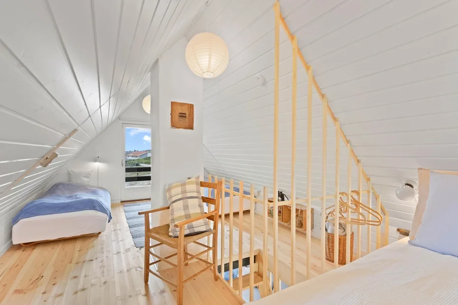 A bright attic bedroom has sloped white ceilings and a wooden stair railing.