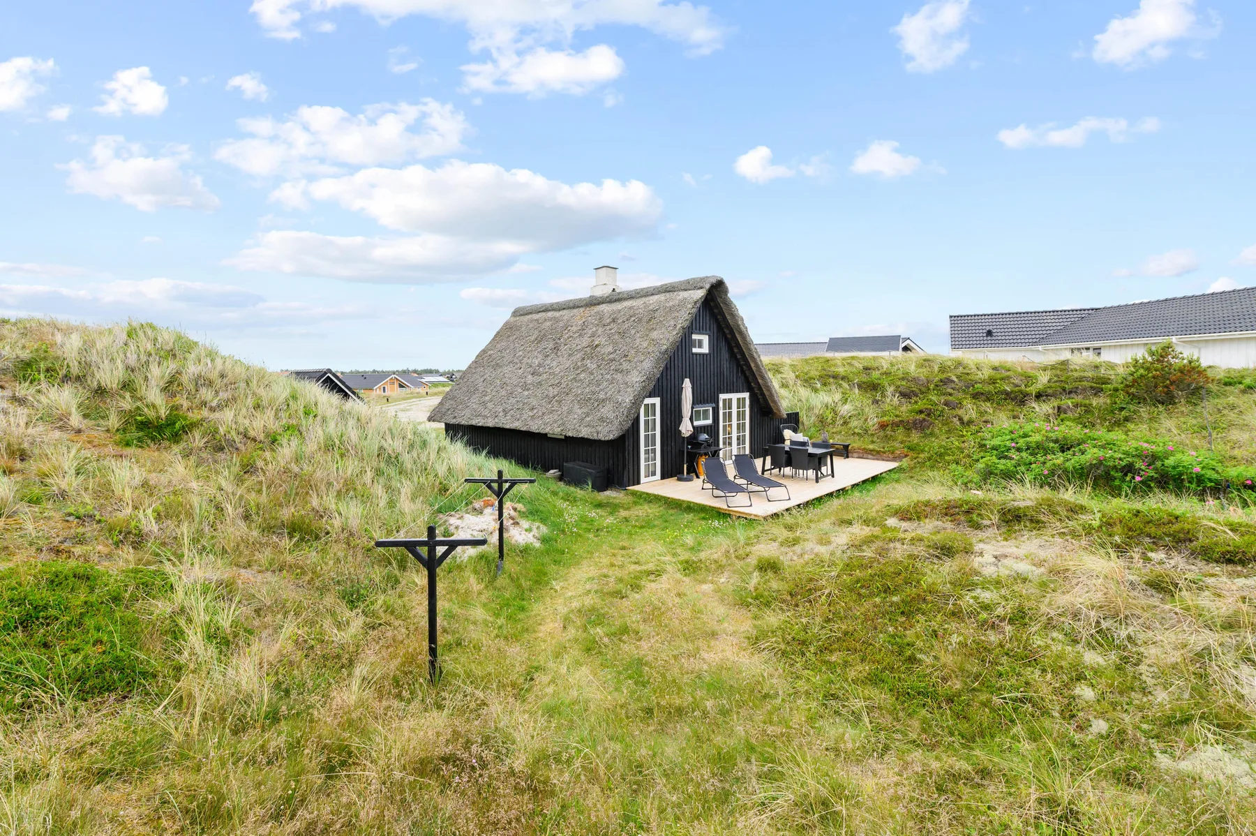 A thatched cottage sits among grassy dunes with outdoor seating by the entrance.