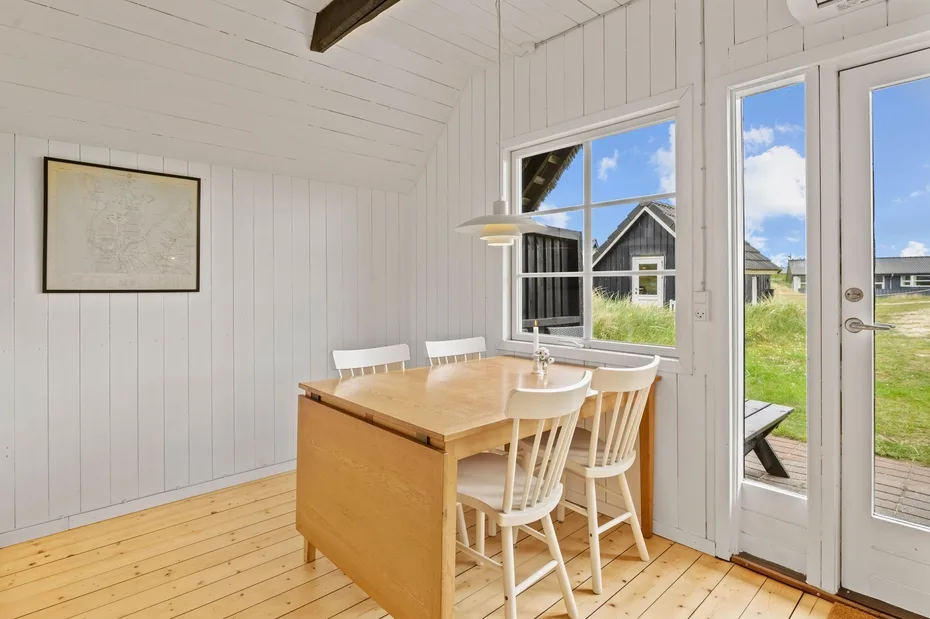 A bright dining area has a wooden table, white chairs, and windows overlooking a grassy yard.