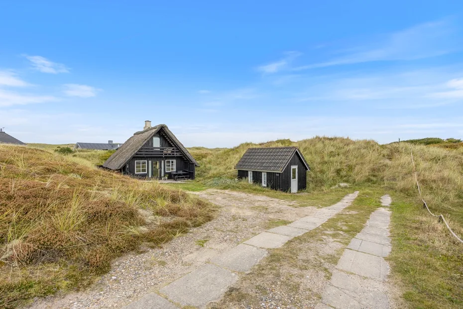 A dark cottage and a small shed sit among grassy dunes beside a paved path.