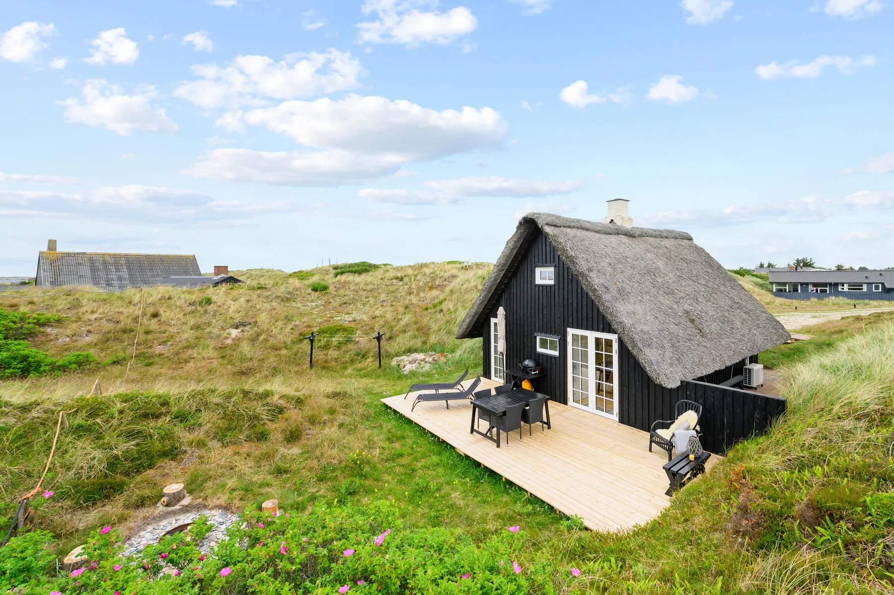 A black cottage sits among grassy dunes, with a wooden deck and outdoor seating.
