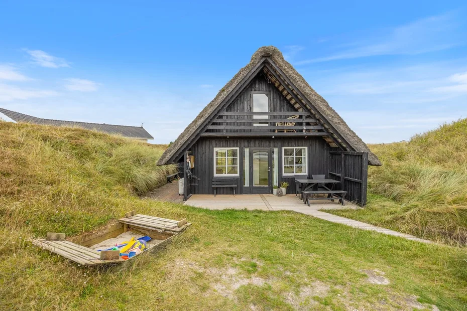 A dark timber cottage sits among grassy dunes beneath a clear sky.