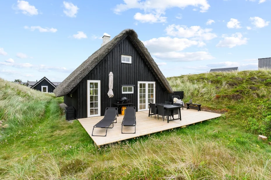 A dark cottage with a steep thatched roof opens onto a deck with outdoor seating.