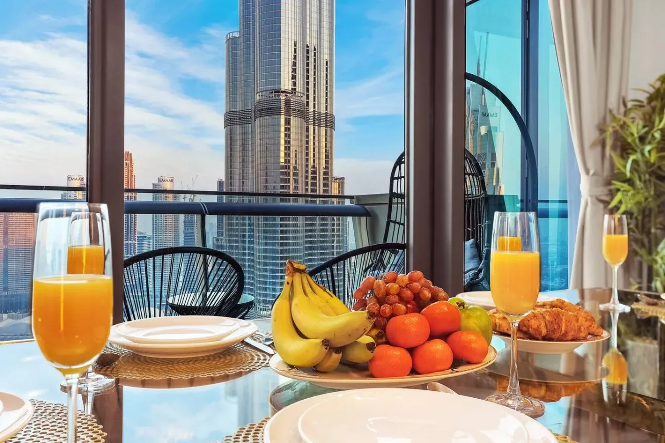 A breakfast table with fruit and juice sits beside windows overlooking city towers.