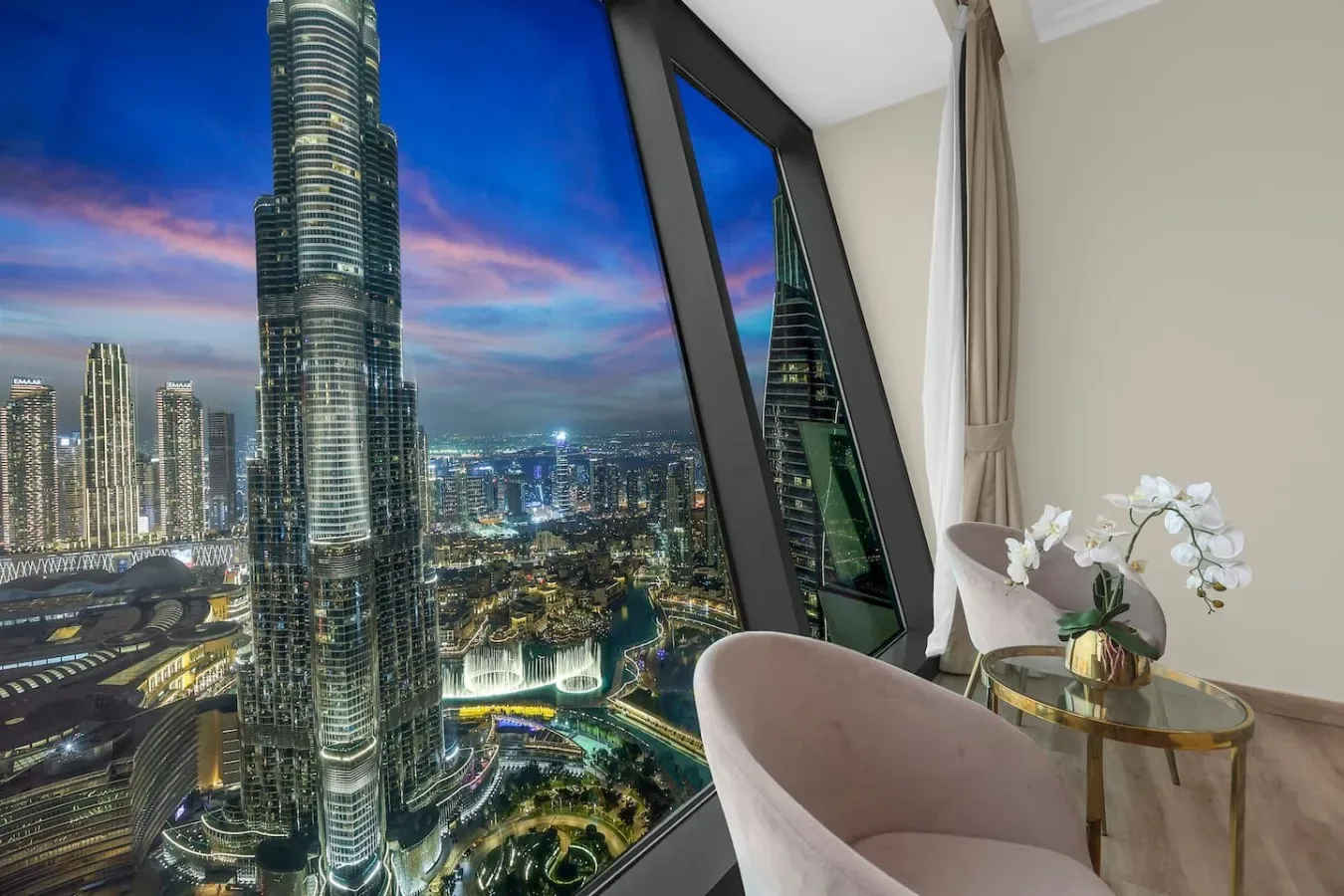 A high-rise room overlooks a brightly lit city skyline at dusk.