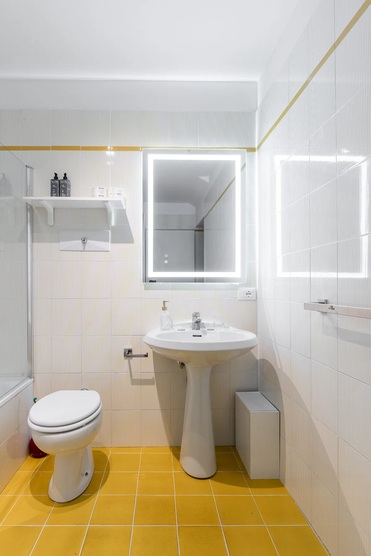 Un baño luminoso tiene un lavabo con pedestal, un espejo de pared y suelo de baldosas amarillas.