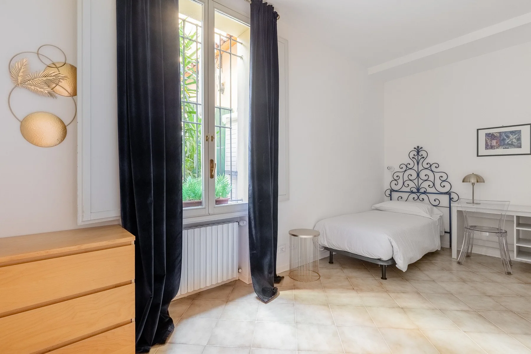 Un dormitorio luminoso con una cama junto a una ventana alta y cortinas oscuras.
