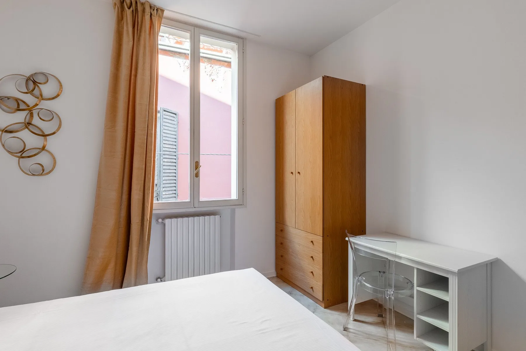 Un dormitorio luminoso con cama, armario de madera y escritorio junto a una ventana.