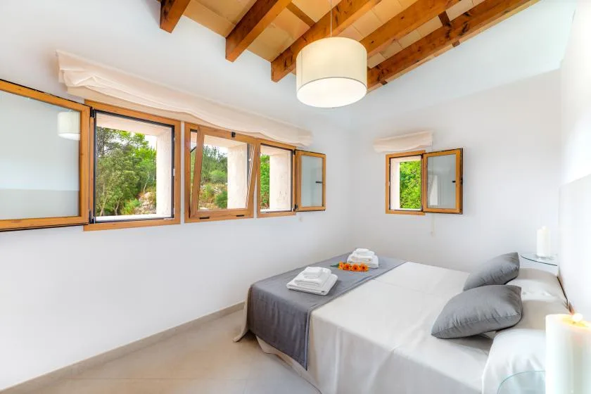 Un dormitorio luminoso con una cama junto a varias ventanas y vigas de madera vistas.