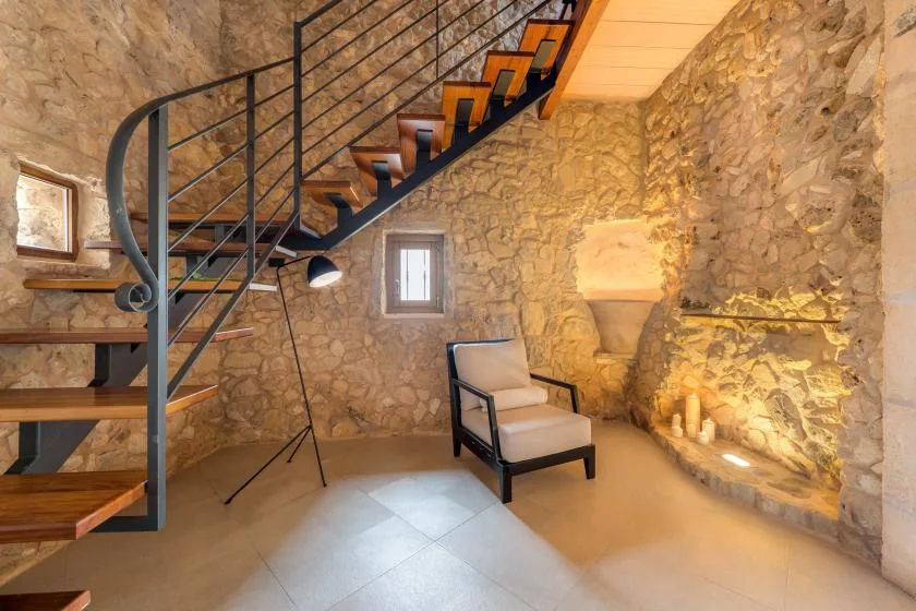 Una zona de estar con paredes de piedra, una escalera y un sillón acolchado.