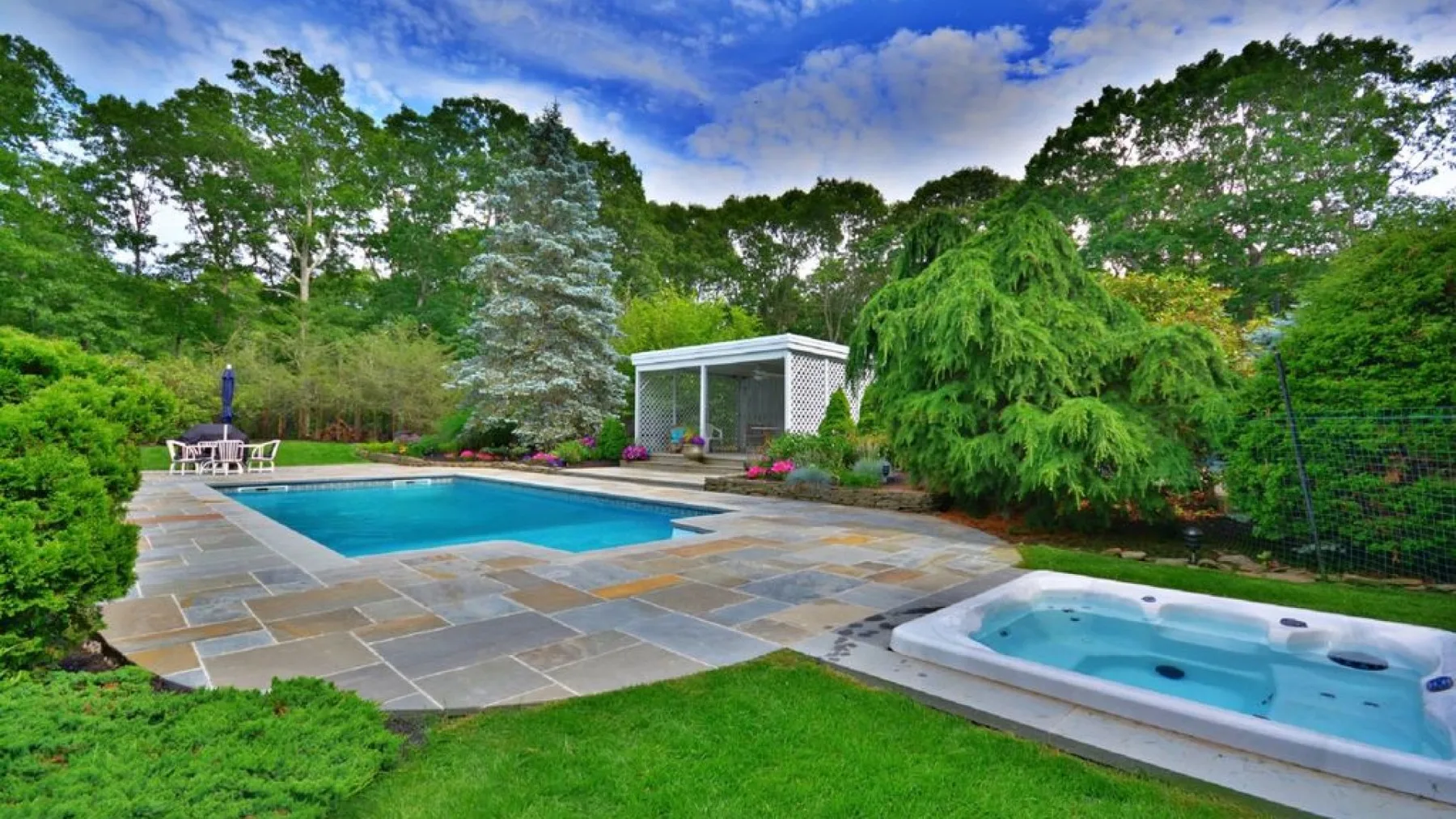 An outdoor pool and hot tub sit beside a stone patio in a landscaped garden.