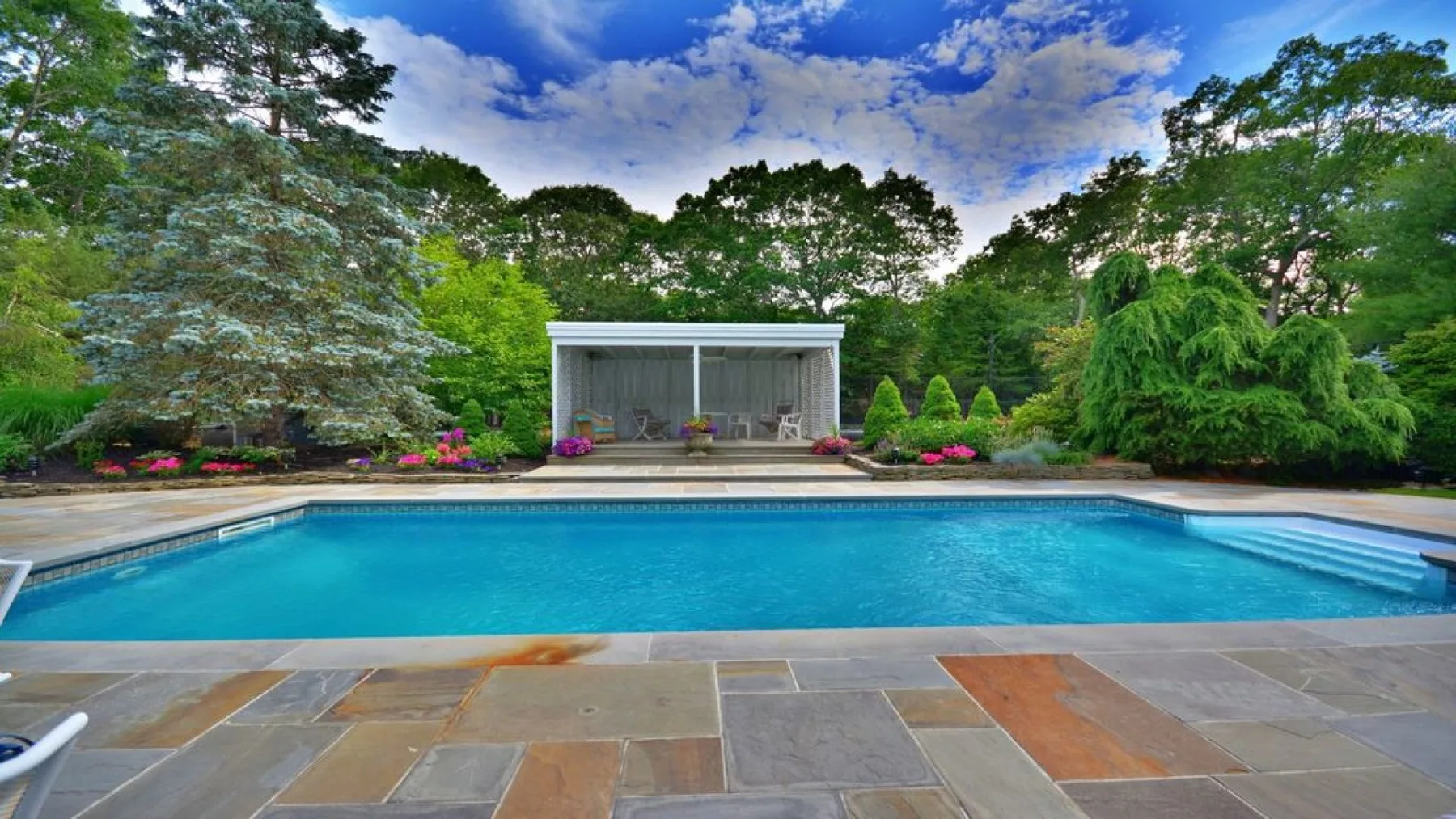An outdoor swimming pool sits before a glass-fronted pavilion and landscaped garden.