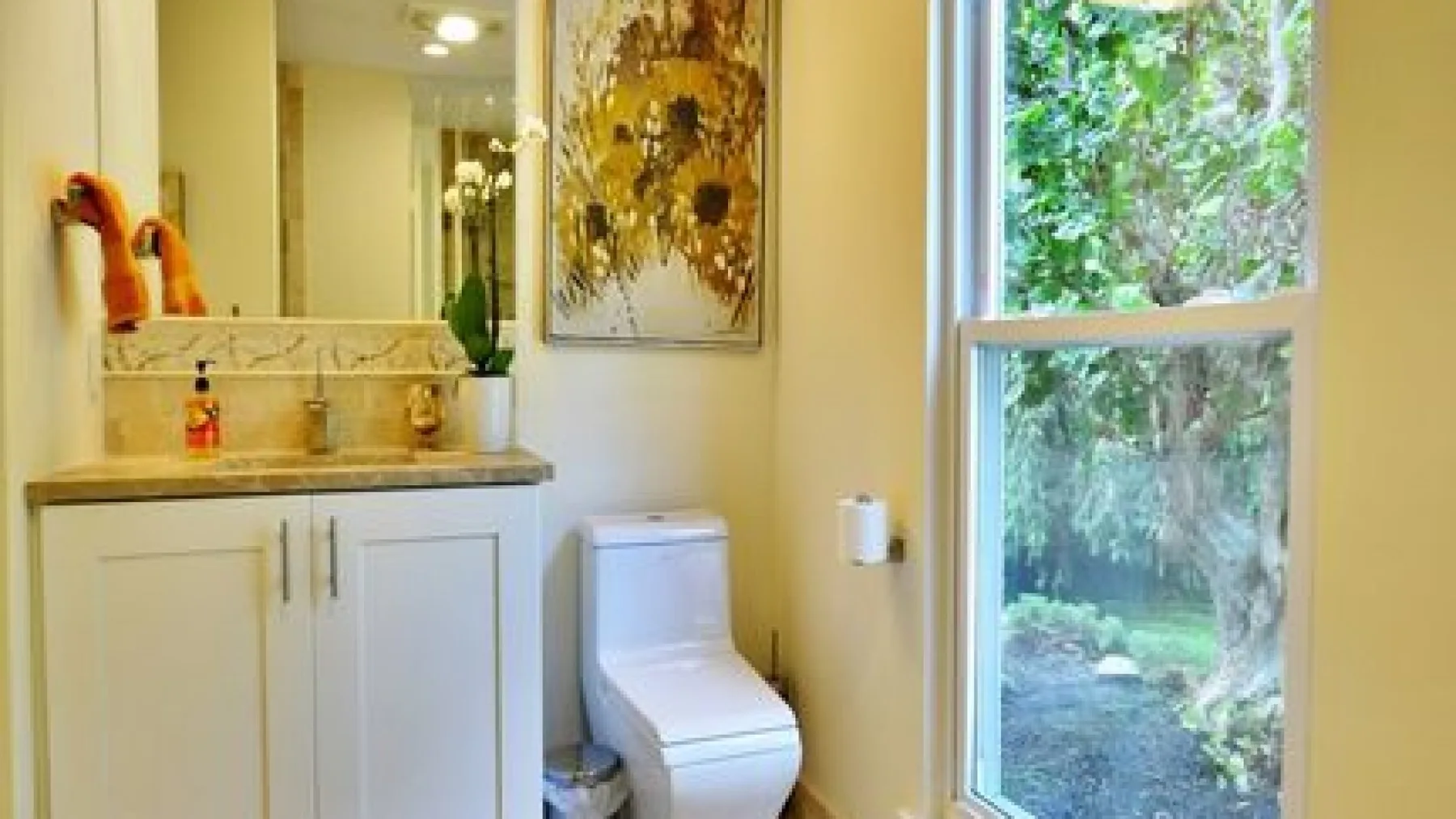 A bathroom with a vanity, toilet, and large window overlooking greenery.