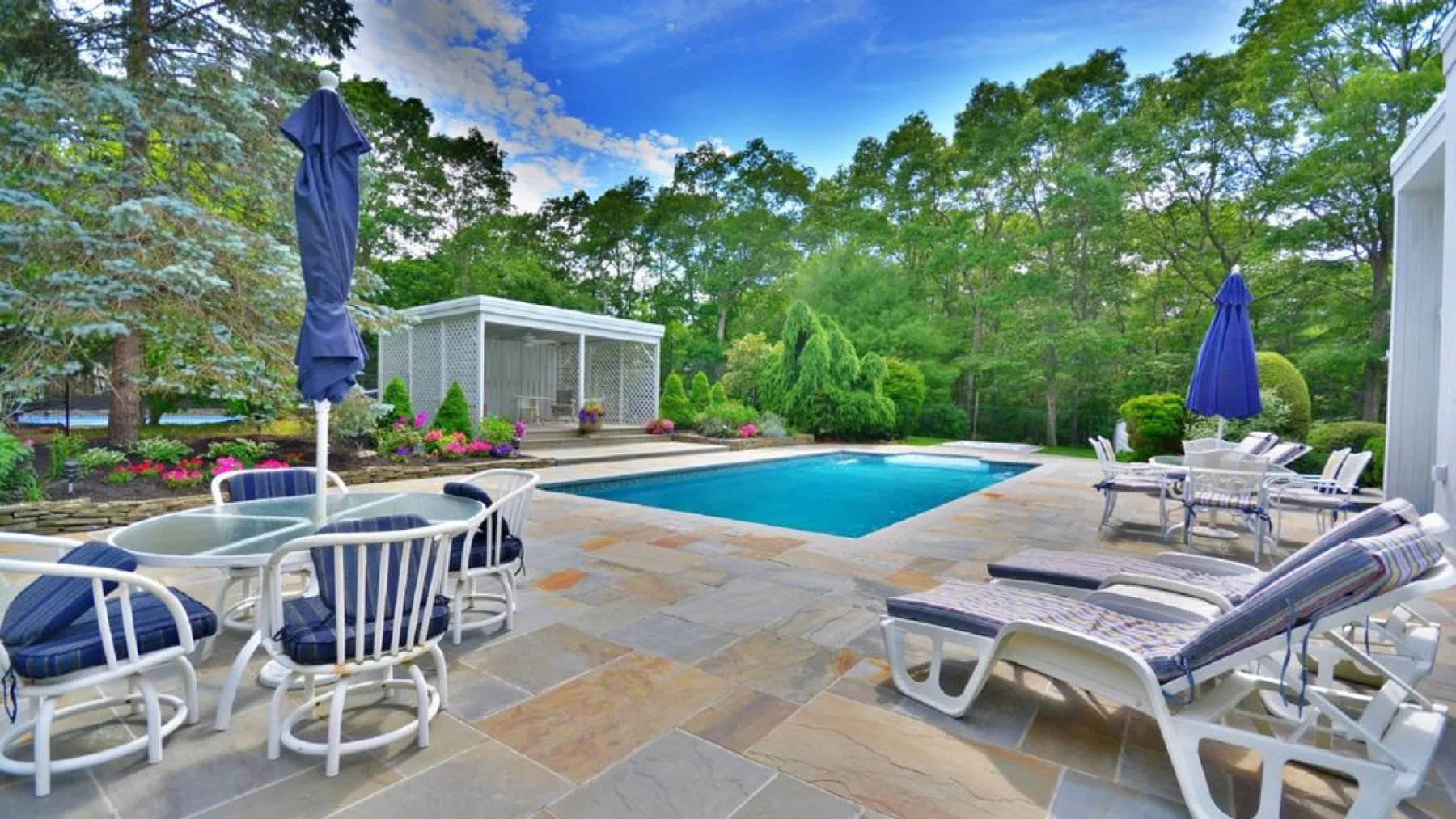 An outdoor pool area with lounge chairs, tables, and surrounding trees.