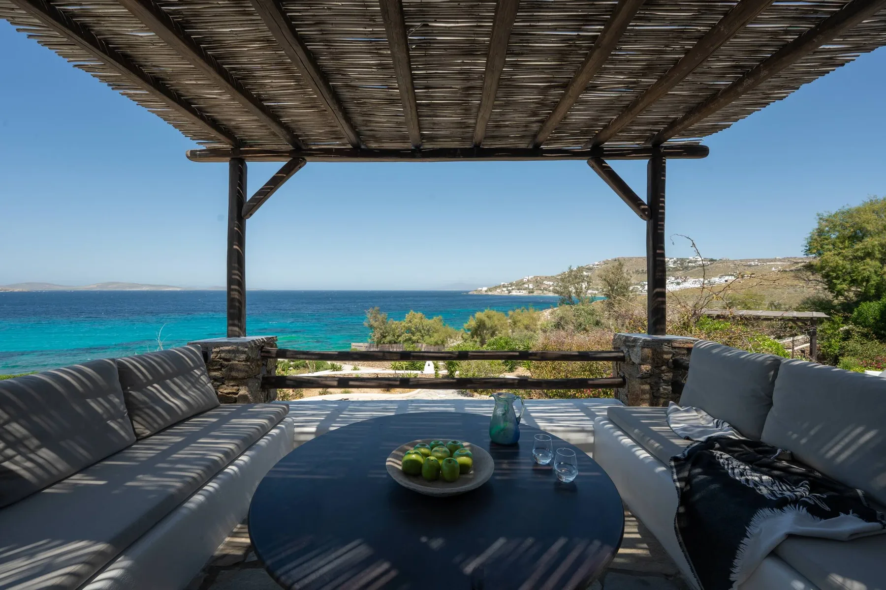 A shaded outdoor seating area overlooks the sea, with a low table in front.