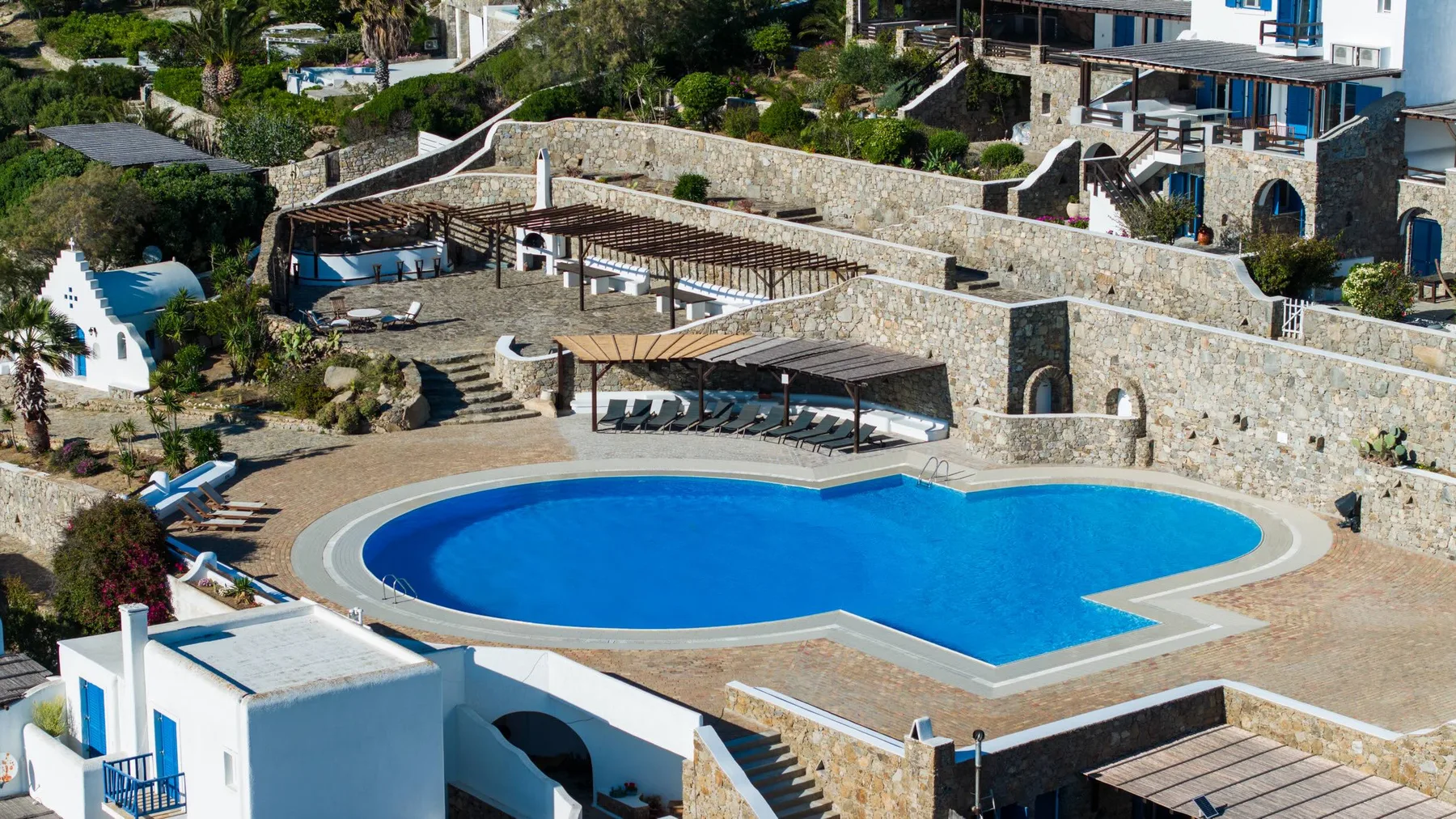 An outdoor swimming pool sits among stone terraces and shaded seating areas.