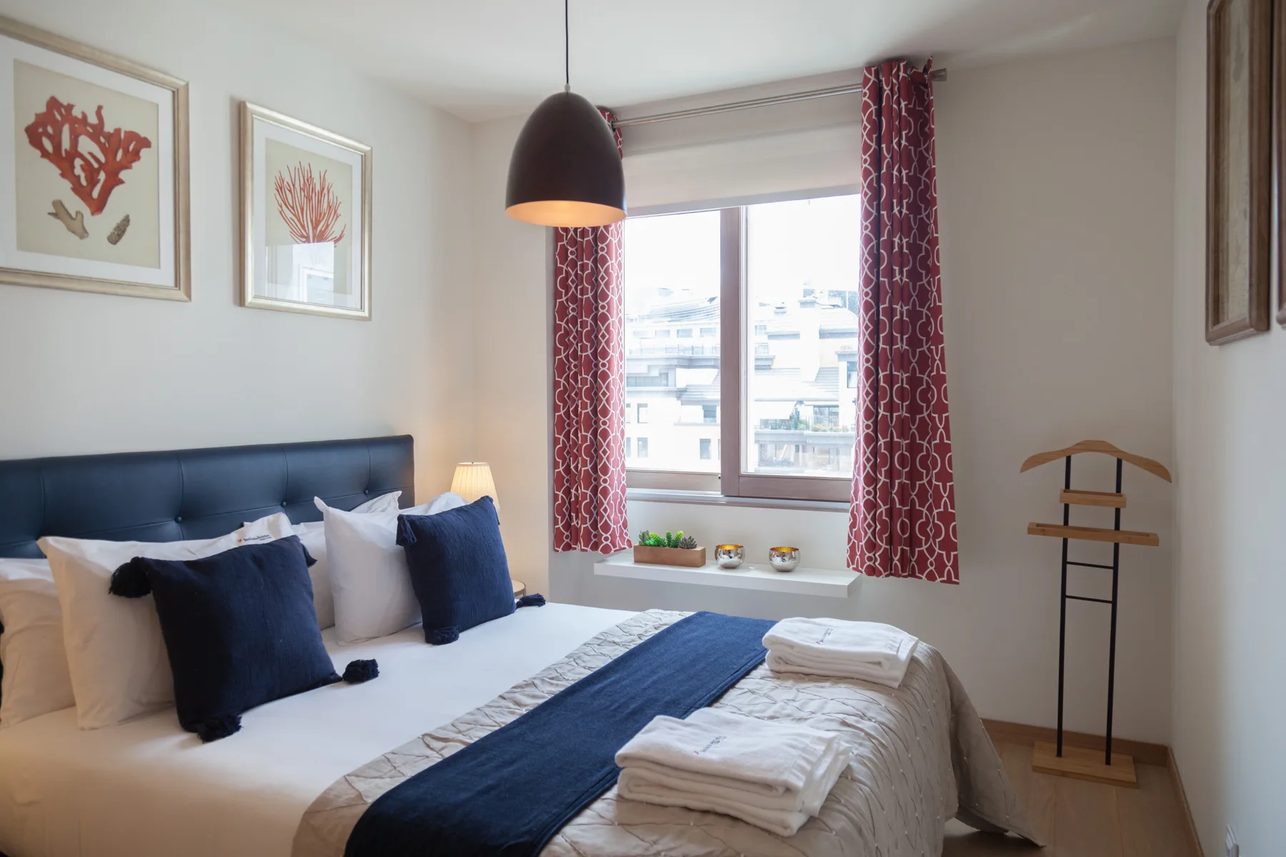 Un dormitorio con una cama bien tendida y una ventana grande con vistas a edificios cercanos.