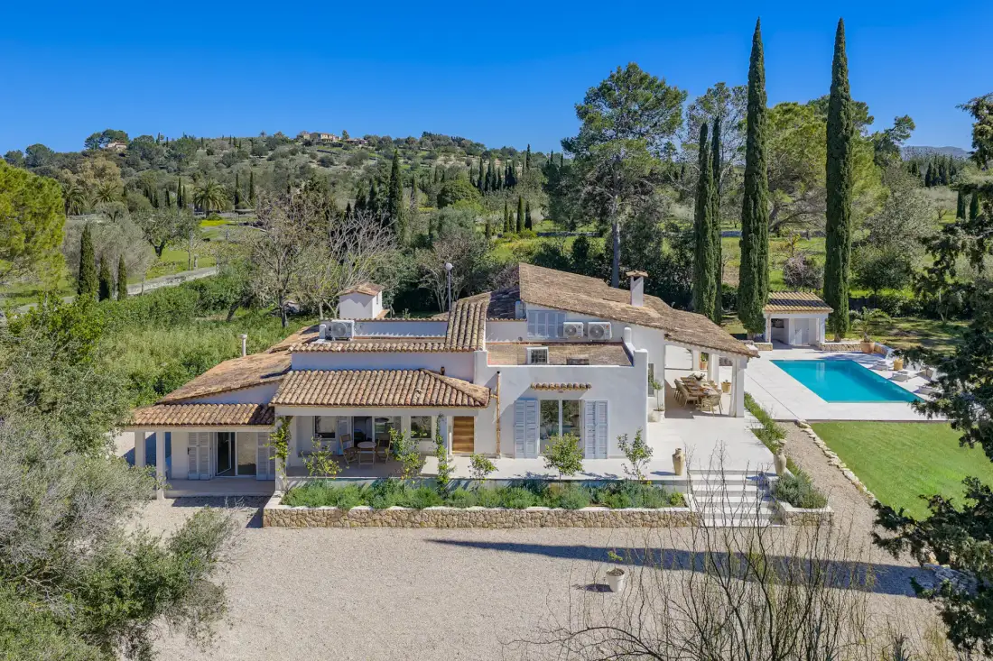 Aerial view of a Mediterranean-style villa with a pool, surrounded by trees and a manicured garden under a clear blue sky.