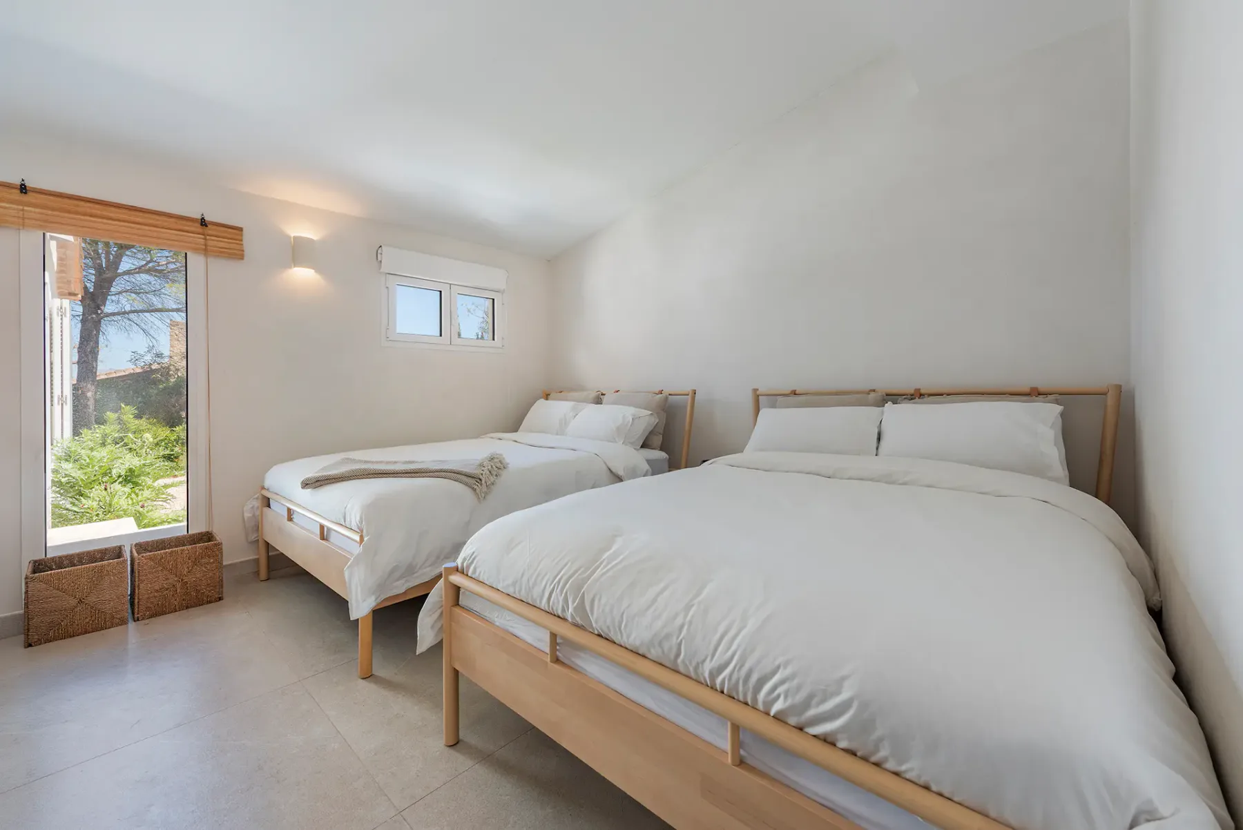 A bright bedroom has two wooden beds with white bedding and a large window overlooking greenery.