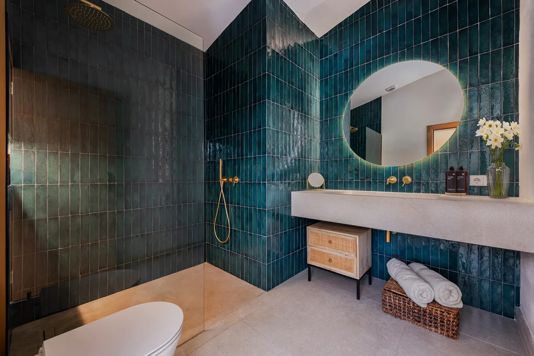 Modern bathroom with dark green tiled walls, round mirror, gold fixtures, minimalistic vanity, shower, and decorative elements like flowers and towels.