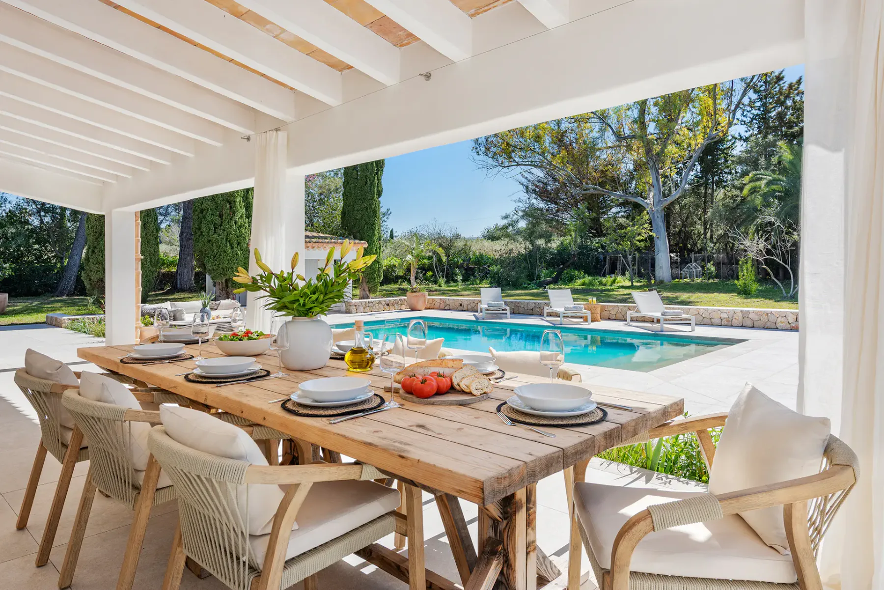 Outdoor dining area with a wooden table set for a meal, overlooking a swimming pool surrounded by lounge chairs and greenery.