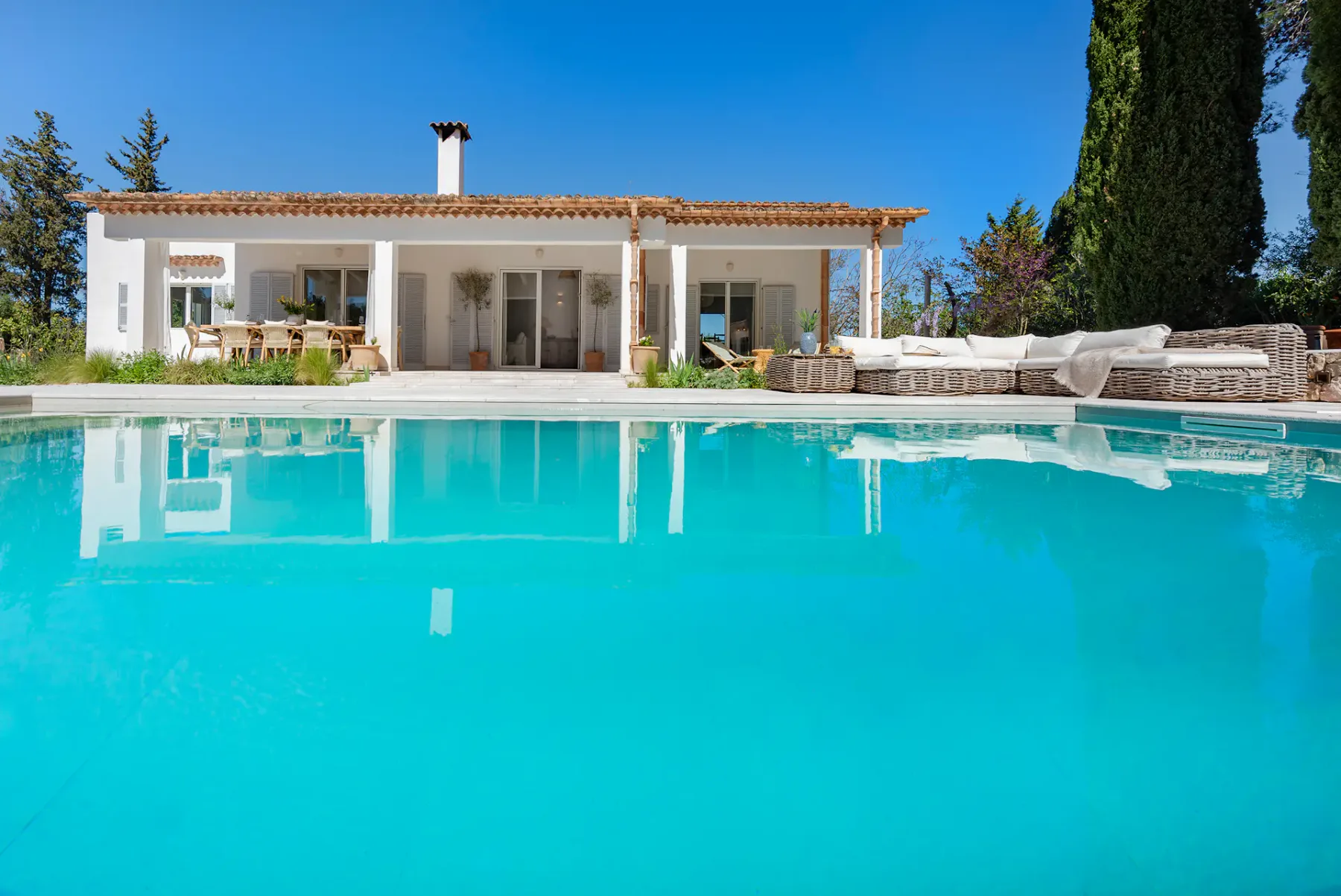 A swimming pool in front of a house, with outdoor seating along the far edge.
