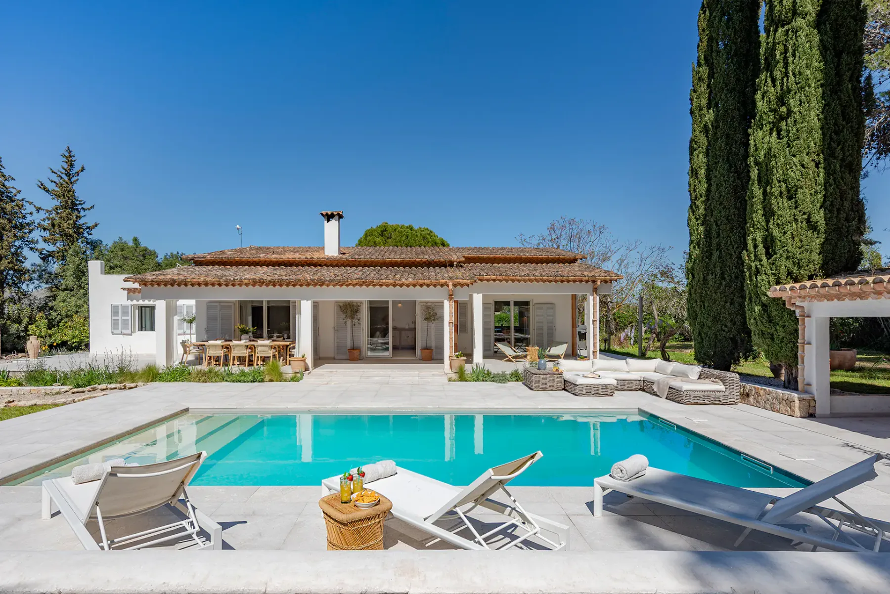 Outdoor pool with four lounge chairs on deck, surrounded by lush trees and greenery under a clear blue sky.