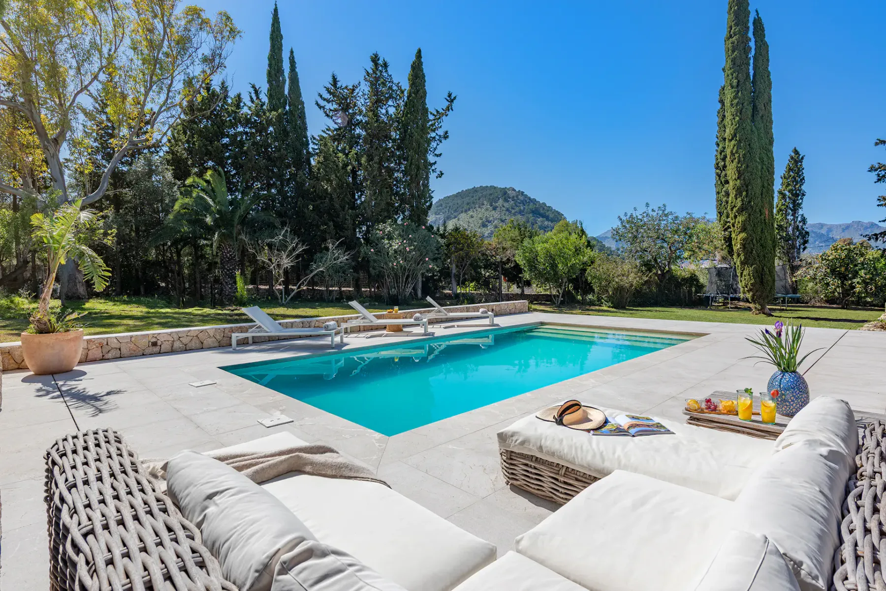 Outdoor pool with four lounge chairs on deck, surrounded by lush trees and greenery under a clear blue sky.