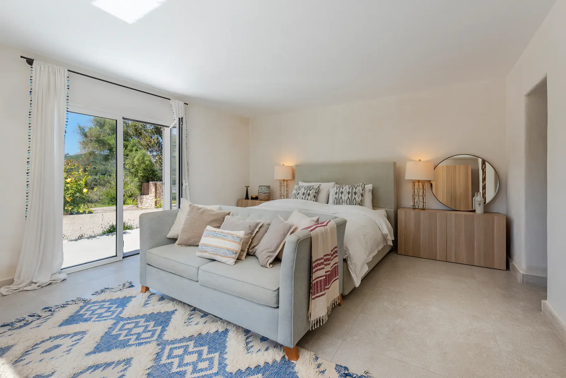 A bright bedroom with a bed, seating, and glass doors overlooking greenery.