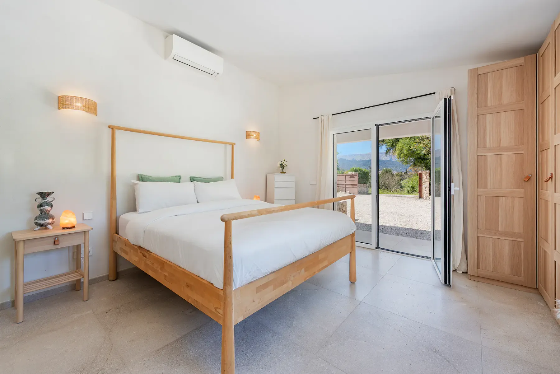 Bright bedroom with a wooden bed, white linens, and open patio doors revealing a scenic outdoor view. Light walls and minimalist decor.