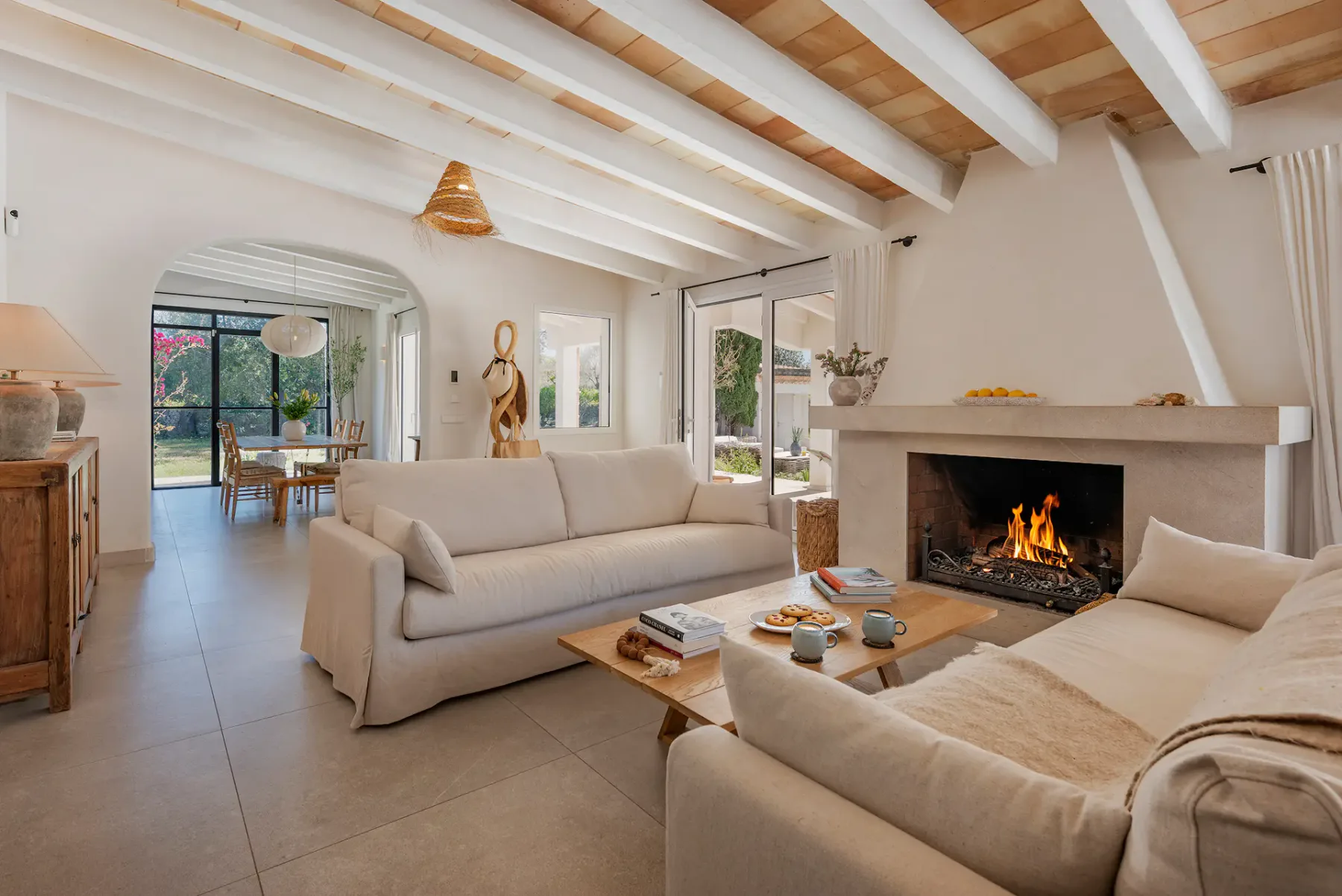 A bright living room has beige seating, a coffee table, and a lit fireplace.