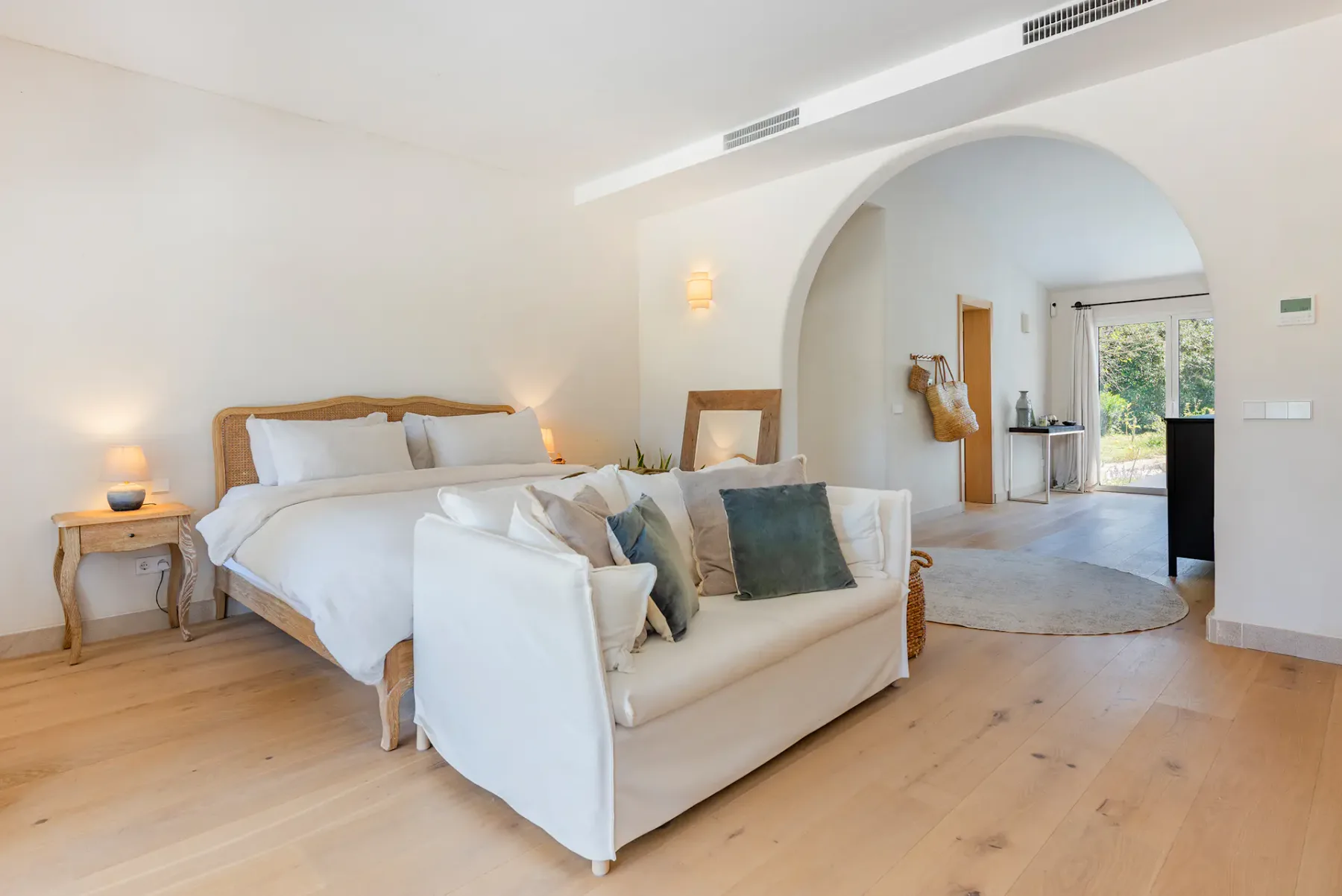 Bright bedroom with a double bed, cozy sofa, wooden floor, and archway leading to a sunlit room with garden view.