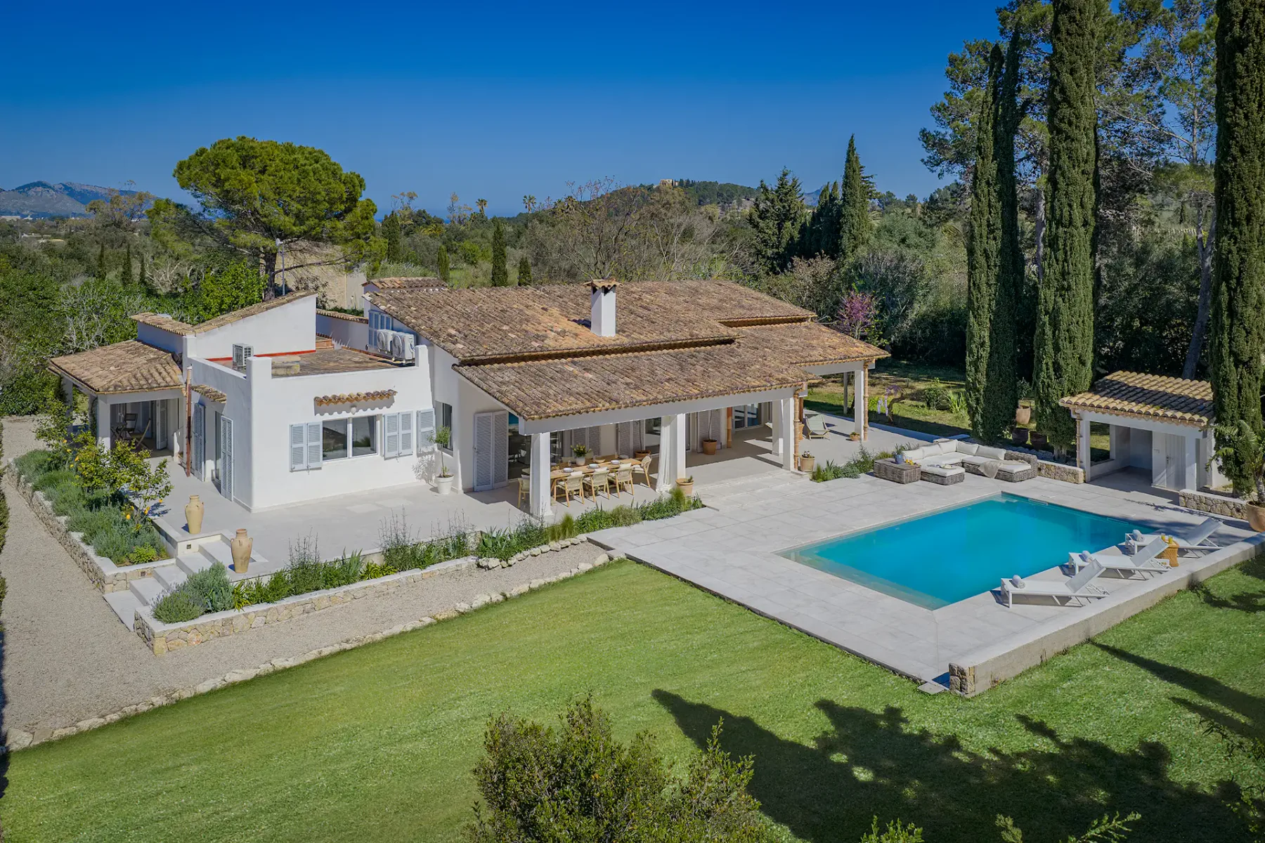 Aerial view of a Mediterranean-style villa with a pool, surrounded by trees and a manicured garden under a clear blue sky.