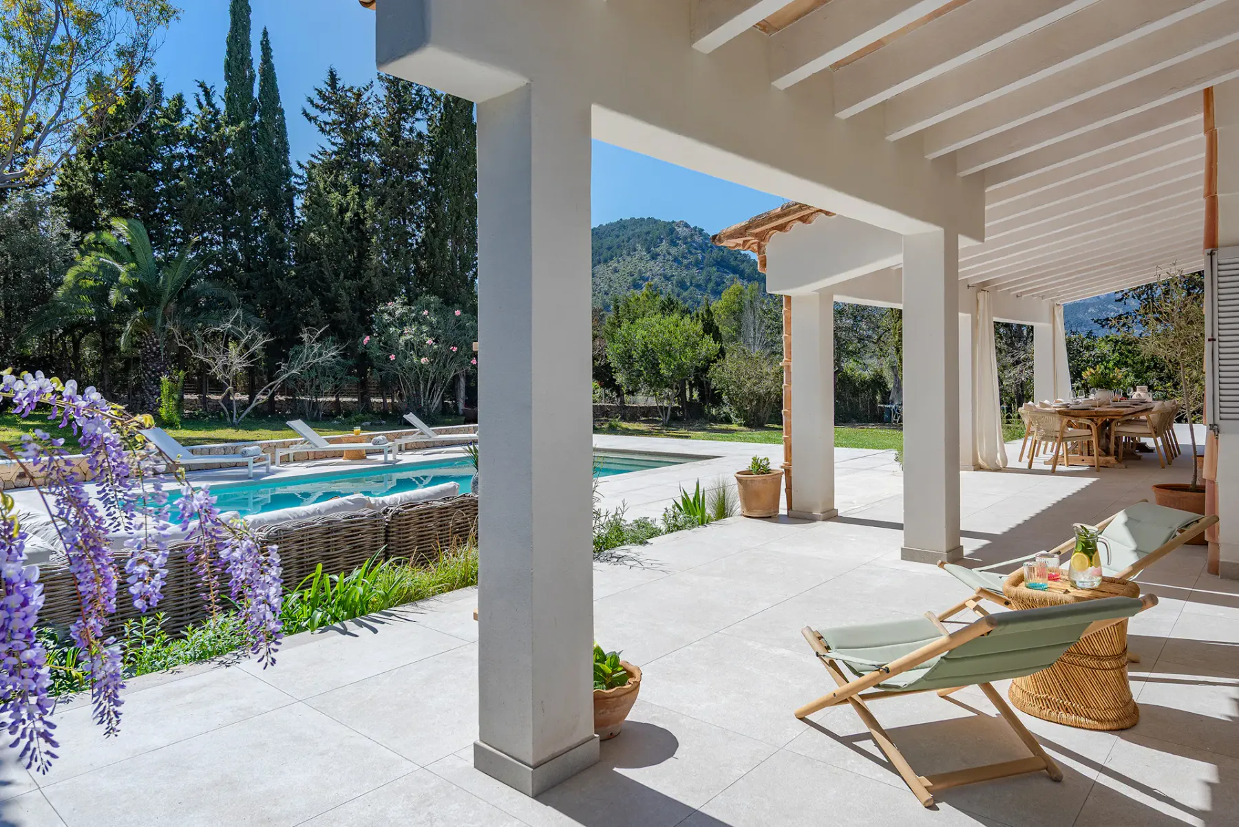 Covered patio with lounge seating beside a swimming pool and surrounding greenery.