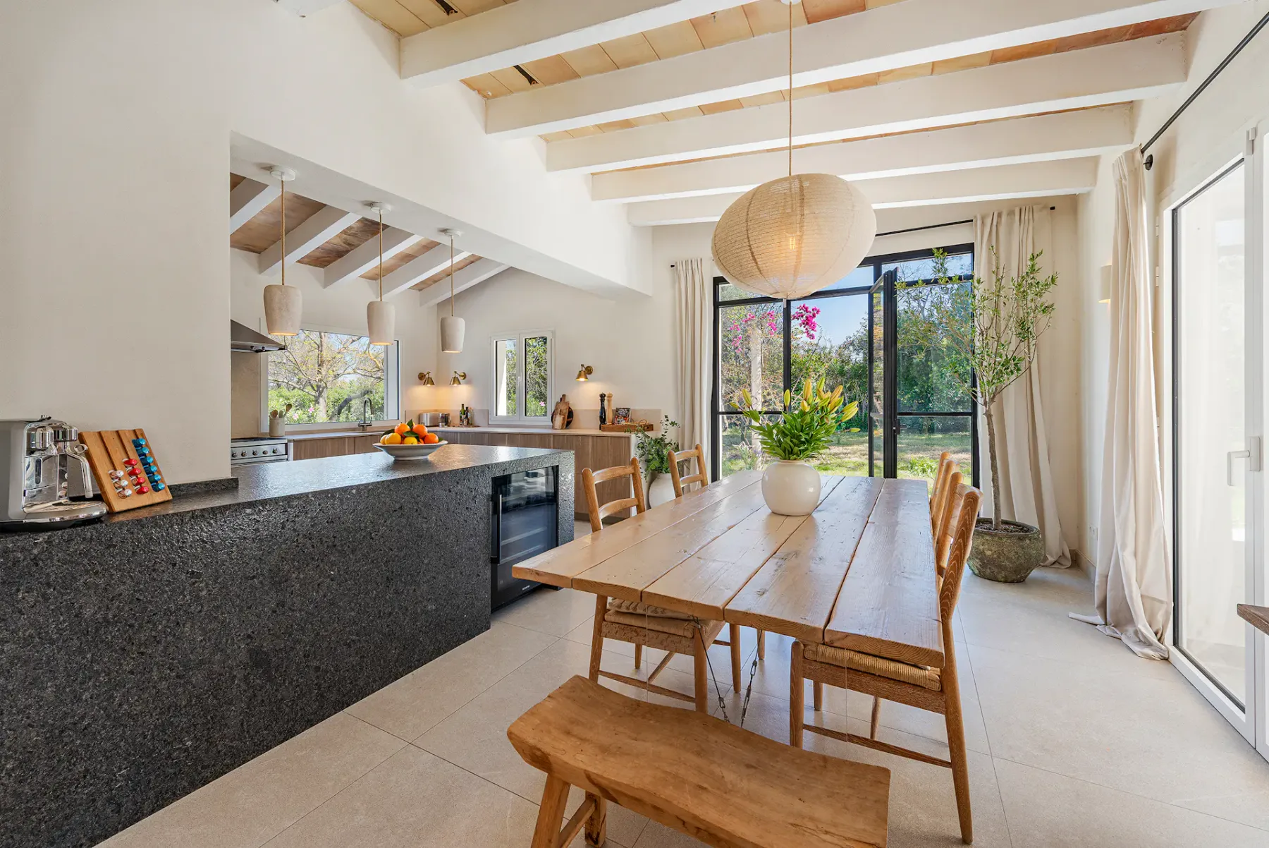 Bright, modern kitchen with wooden table, pendant lights, and light wood cabinetry. Greenery and flowers add a fresh touch.