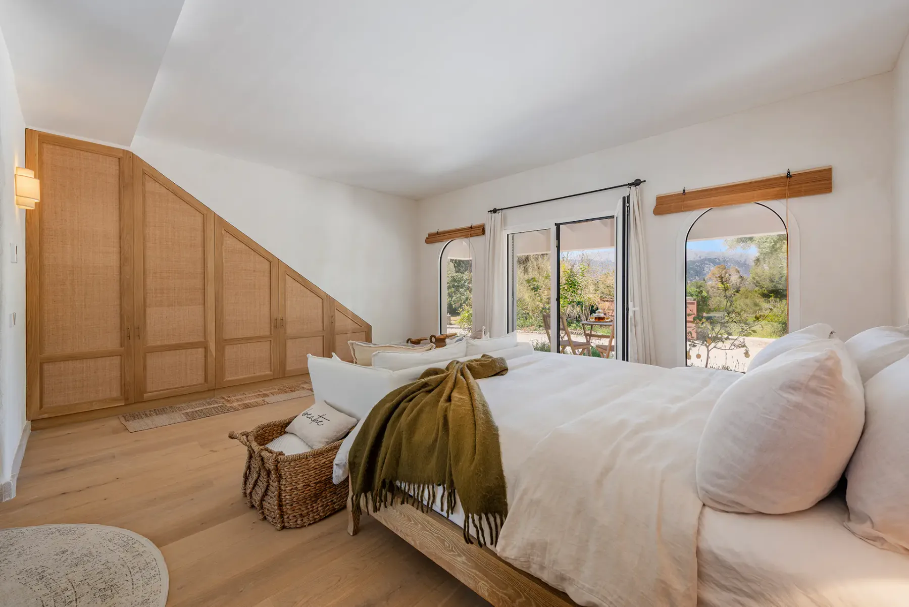 A bright bedroom with a large bed, a woven basket, and windows overlooking greenery.