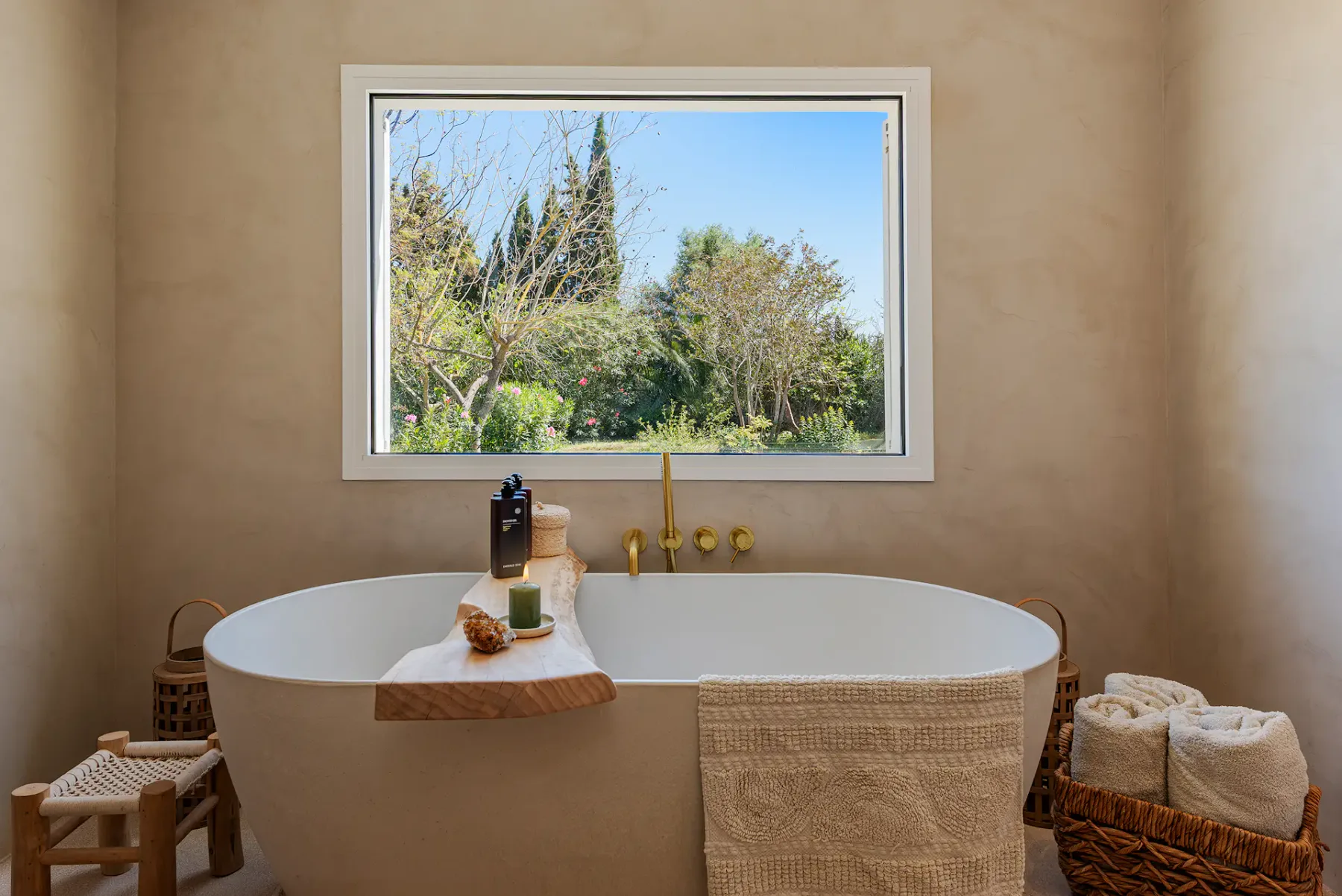 A bathroom with a freestanding tub beneath a window overlooking greenery.