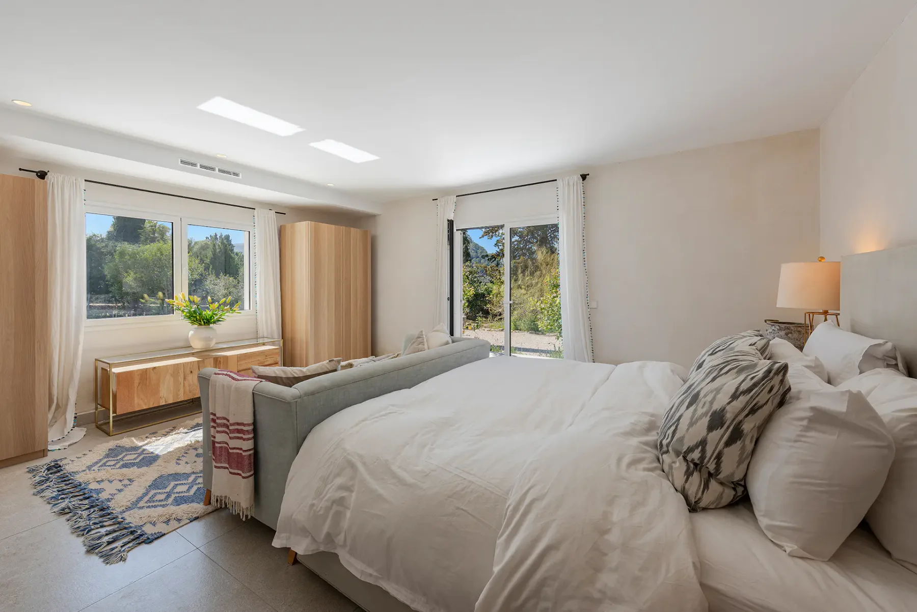 A bedroom with a large bed, seating, and windows overlooking greenery.