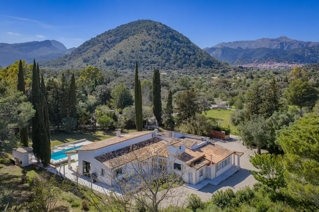 Aerial view of a Mediterranean-style villa with a pool, surrounded by trees and a manicured garden under a clear blue sky.