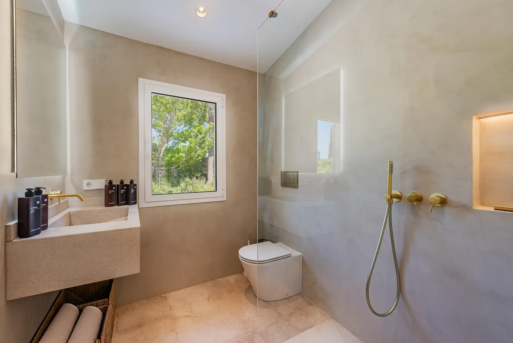 Modern bathroom with beige tones, featuring a wall-mounted toilet, rain shower, sink, window with greenery, and minimalist decor.