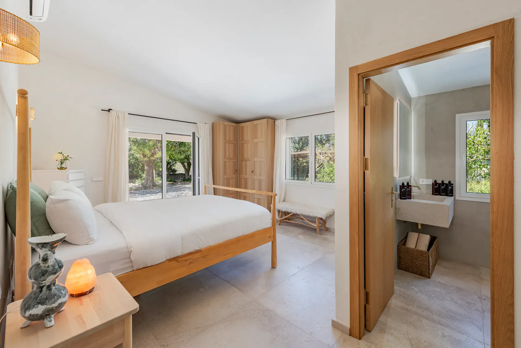 Bright bedroom with a wooden bed, white linens, and open patio doors revealing a scenic outdoor view. Light walls and minimalist decor.