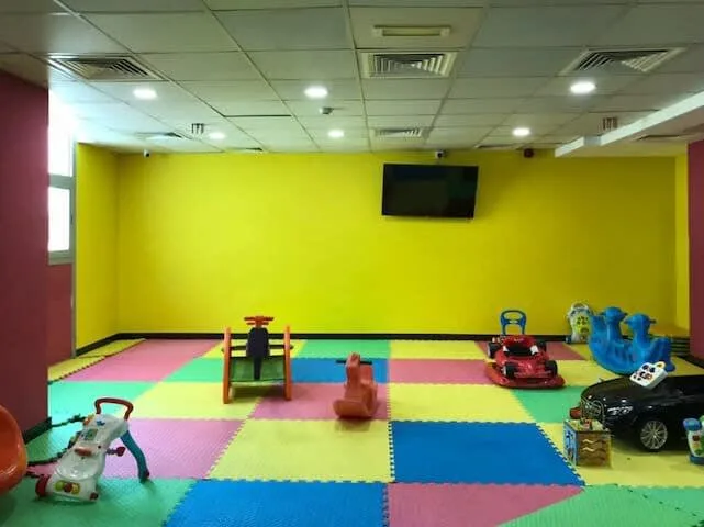 A colorful indoor playroom has padded floor tiles and several children’s toys.
