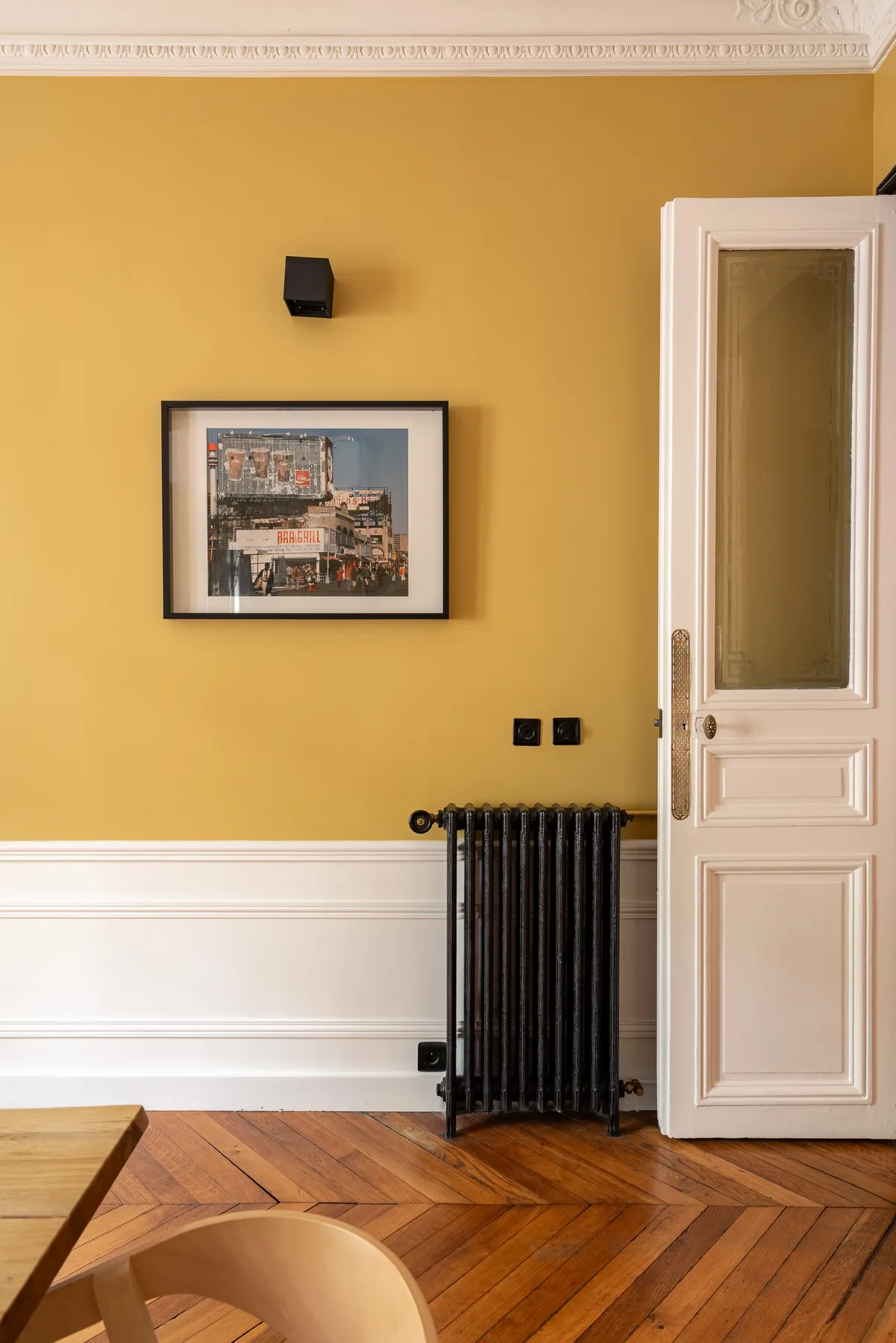 Una habitación con pared amarillo mostaza, una obra enmarcada y una puerta blanca.