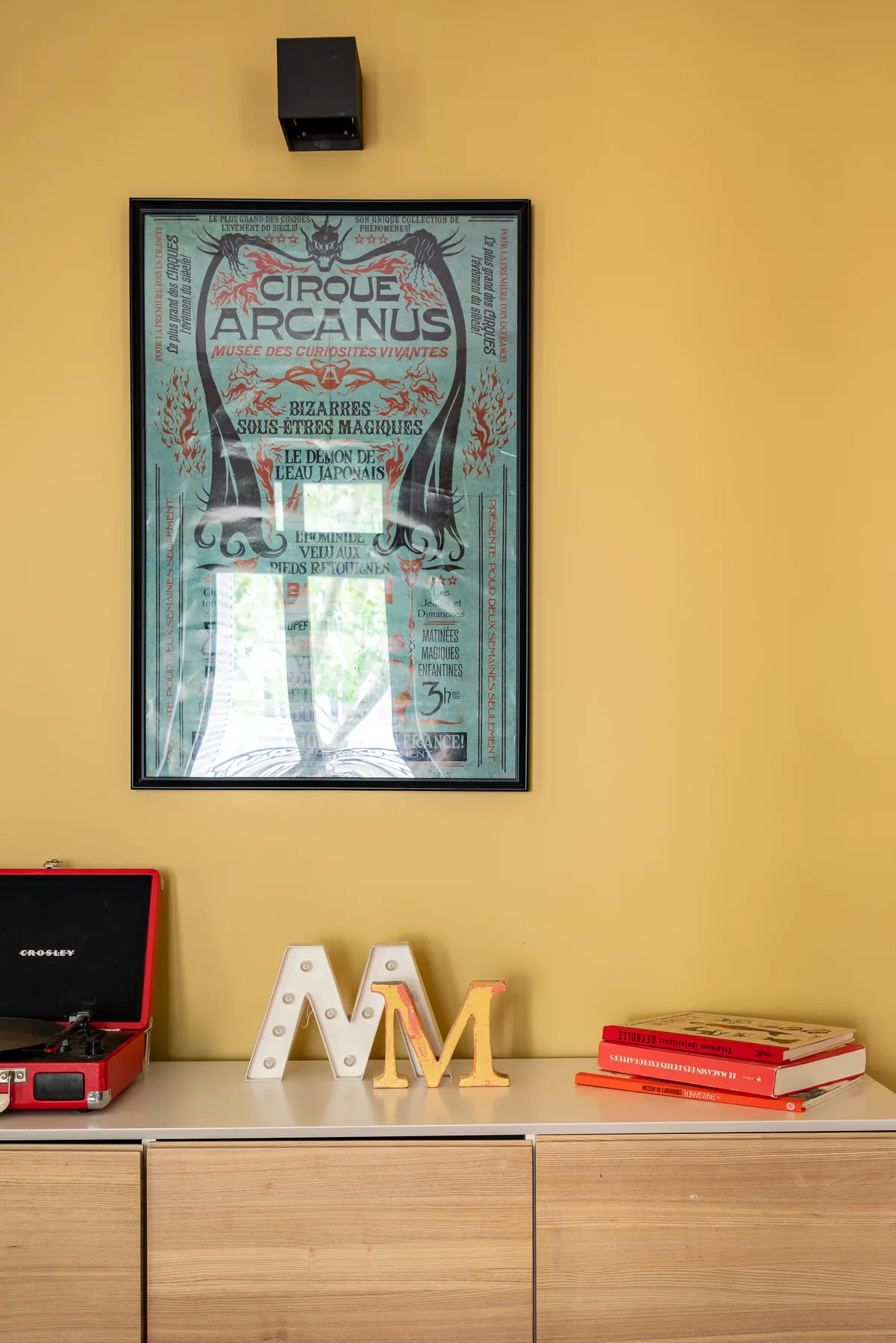 Un cartel de circo enmarcado cuelga sobre un mueble de madera con letras decorativas y libros.