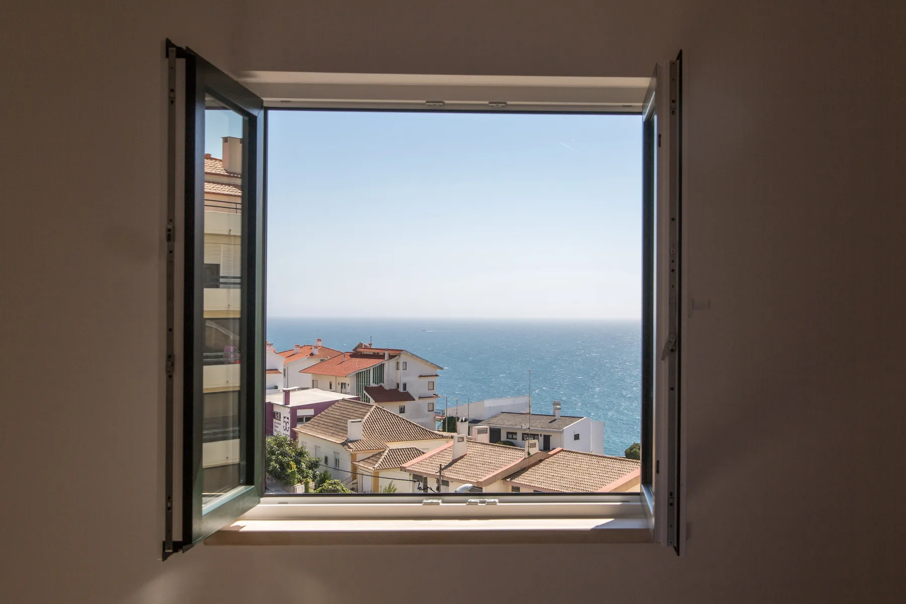 Una ventana abierta enmarca tejados cercanos y una vista del mar.