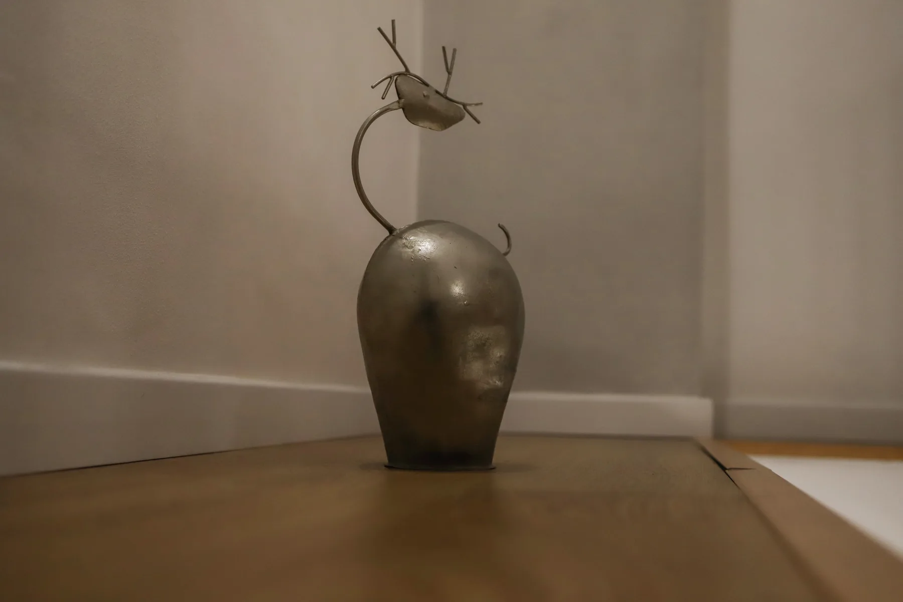 Un jarrón decorativo de metal con un tallo curvo está en el suelo junto a una pared.