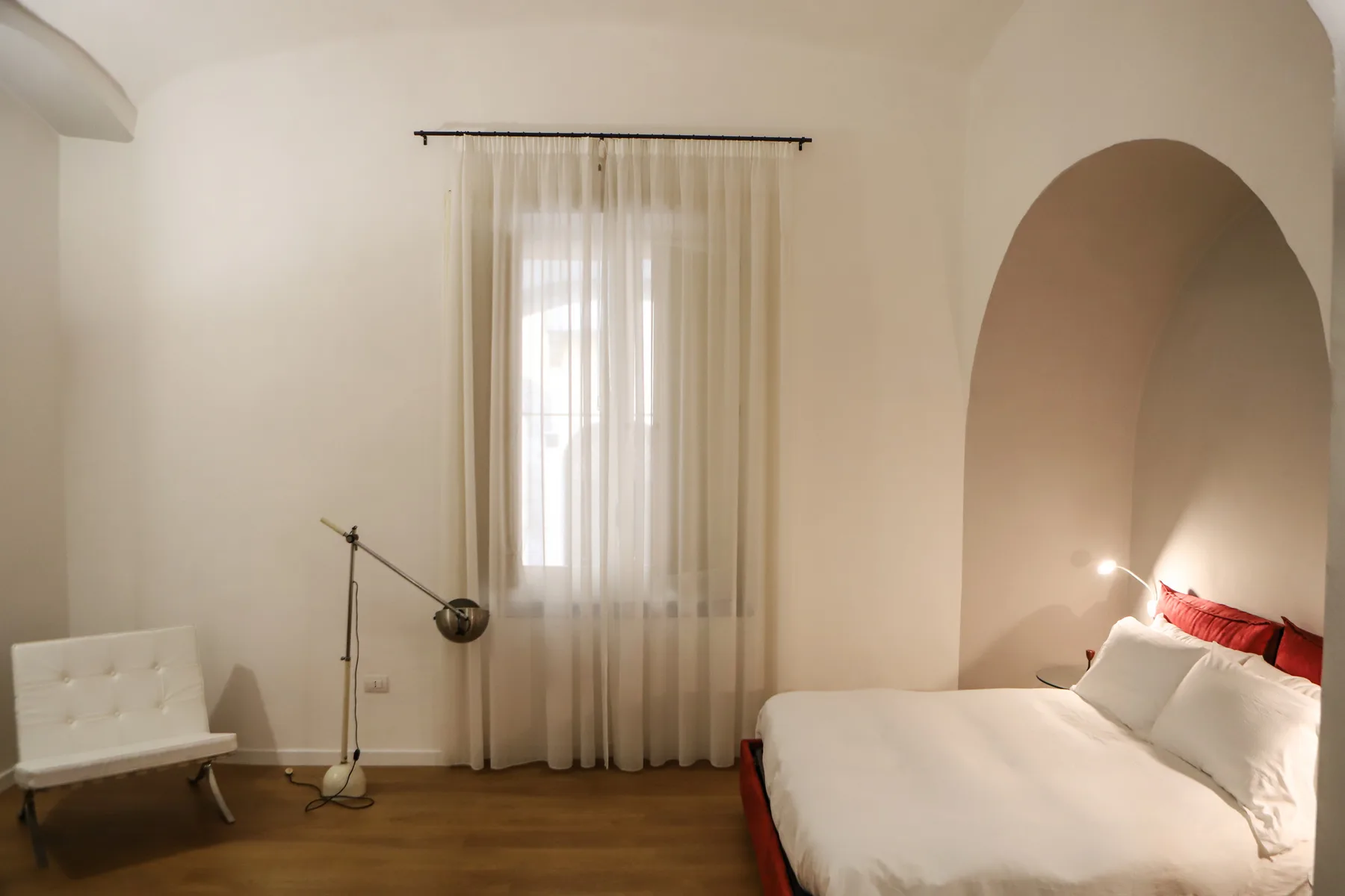 Un dormitorio con una cama junto a un nicho alto en arco y una ventana con cortinas.