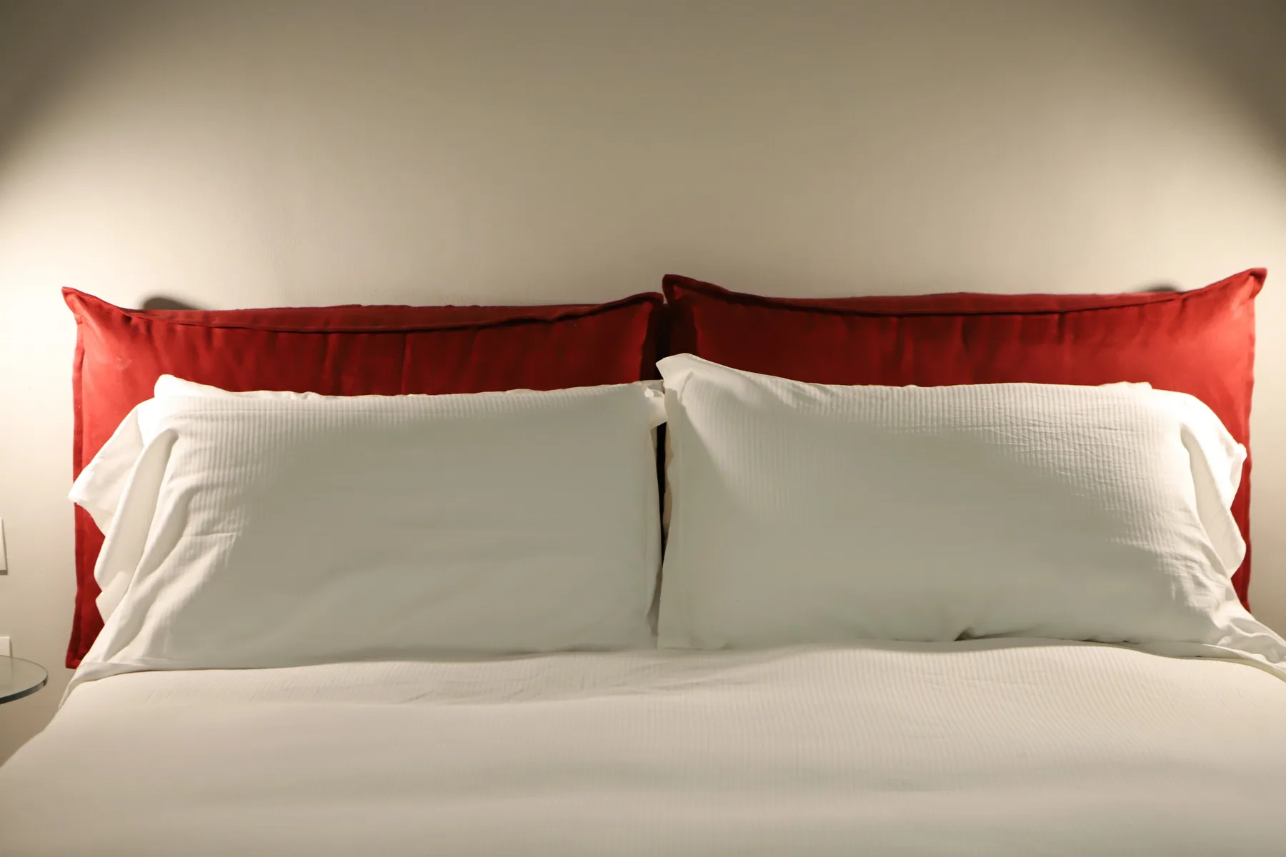 Una cama bien hecha con almohadas blancas y cojines rojos frente a una pared lisa.