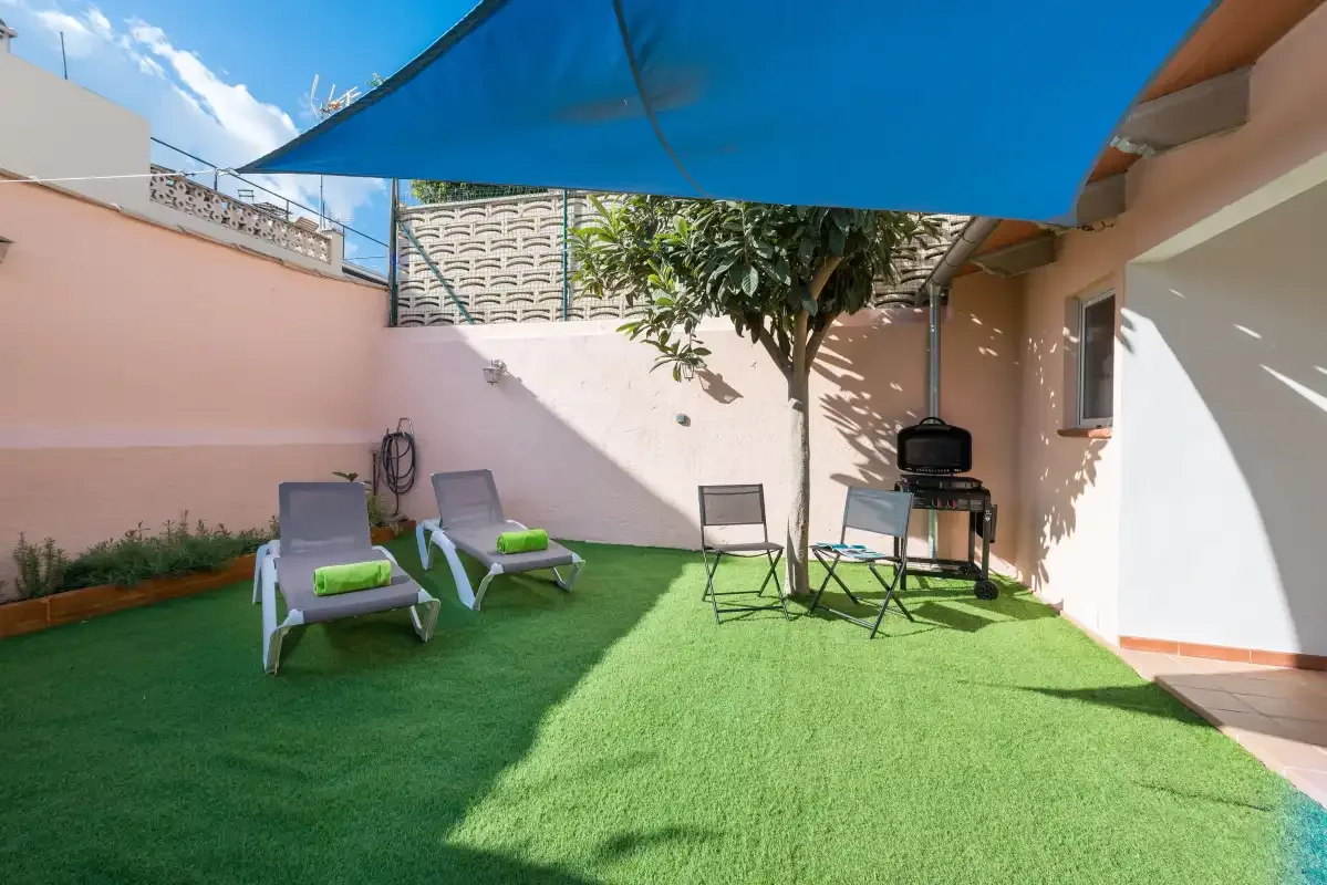 A small outdoor patio with seating, a tree, and a grill under a blue shade canopy.