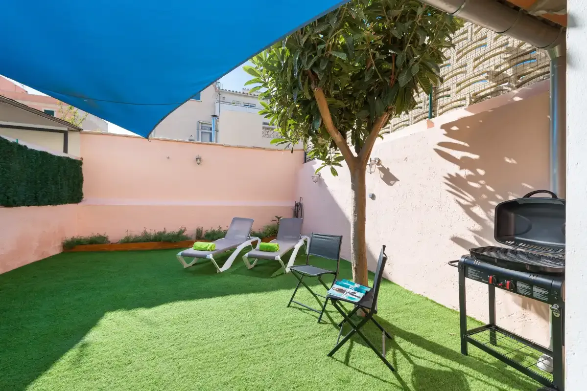 A small outdoor patio has artificial grass, seating, and a barbecue beneath a shade canopy.